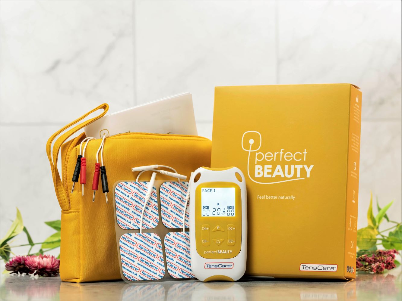A yellow TensCare Perfect Beauty TENS Machine for Wrinkles and Body Toning, including electrodes, wires, a zippered pouch, packaging, and a bonus free set of face pads, is displayed on a marble surface with flowers and leaves in the background.
