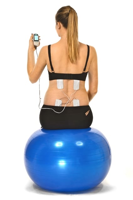 Woman sitting on blue exercise ball with four electrode pads attached to her back, holding a handheld device connected to the electrodes.