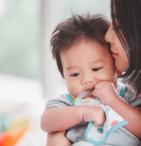 A woman holds a baby who is chewing on a teething toy. Both are indoors, and the background is softly blurred.