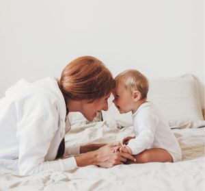Woman and baby sitting on a bed, facing each other and touching foreheads whilst smiling. Both are wearing white clothing in a bright, neutral room.