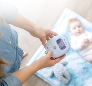 A person holds a portable breast pump with a digital display, whilst a baby lies on a blue blanket in the background.